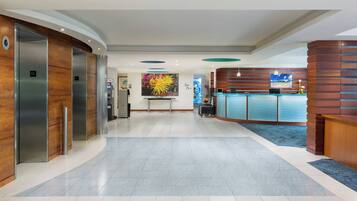 A hotel lobby with a reception desk, elevators, and a large painting on the wall.