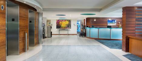 A hotel lobby with a reception desk, elevators, and a large painting on the wall.