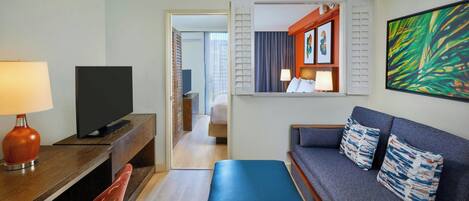 A hotel room with a TV, a desk with a lamp, a sofa with patterned cushions, and a large painting on the wall.
