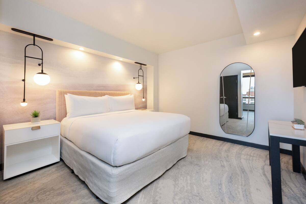 A modern hotel room with a large bed, bedside tables, a mirror, and a small table with a plant.