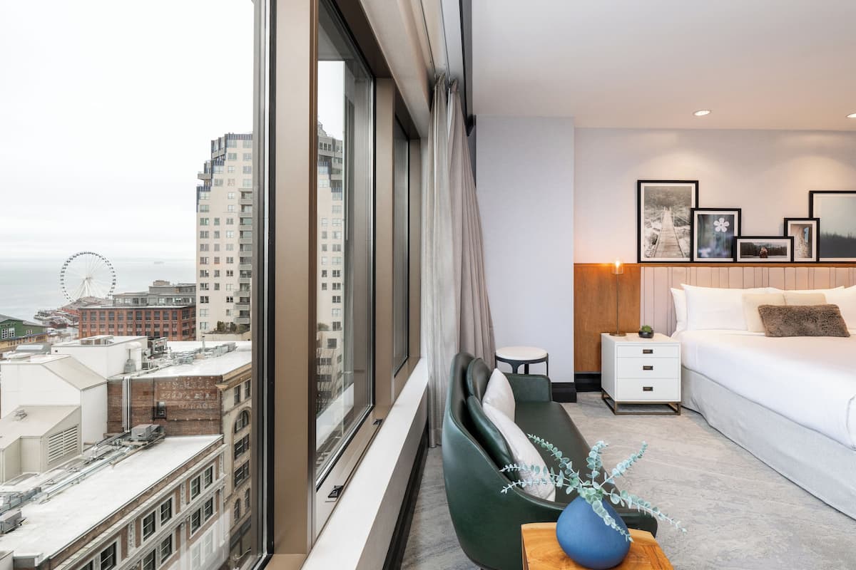 A modern hotel room with a large window, a bed, a nightstand, a chair, and a vase with a plant.