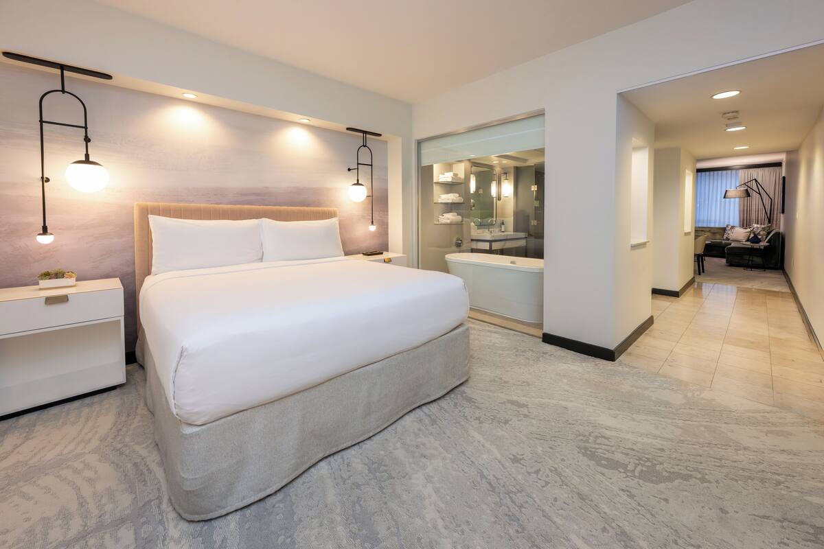 A modern hotel room with a large bed, bedside tables, a glass-enclosed bathroom, and a view of the living area.