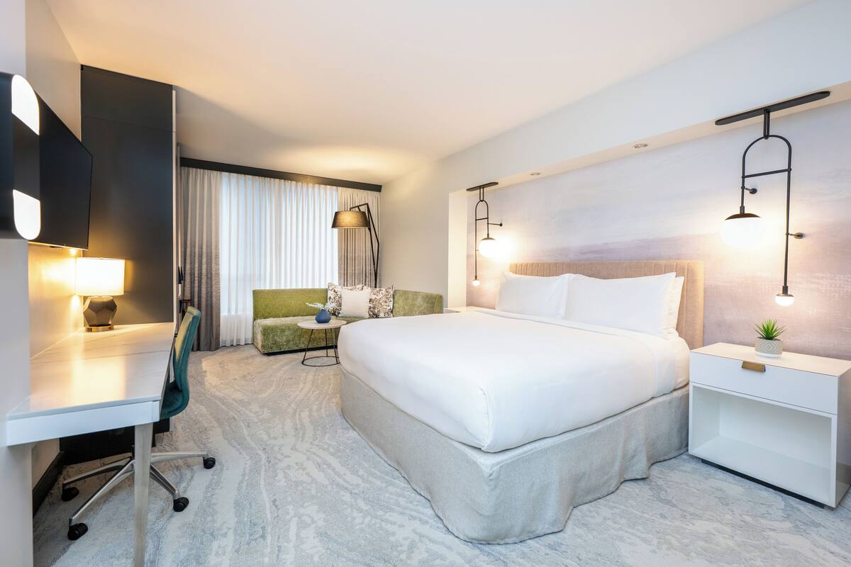 A hotel room with a bed, desk, chair, bedside table, lamp, and a view of the city through the window.