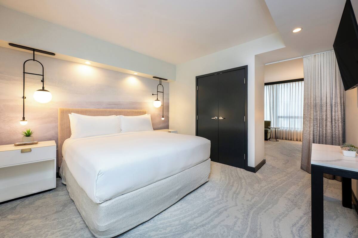A modern hotel room with a large bed, bedside tables, a flat-screen TV, and a door leading to a bathroom.