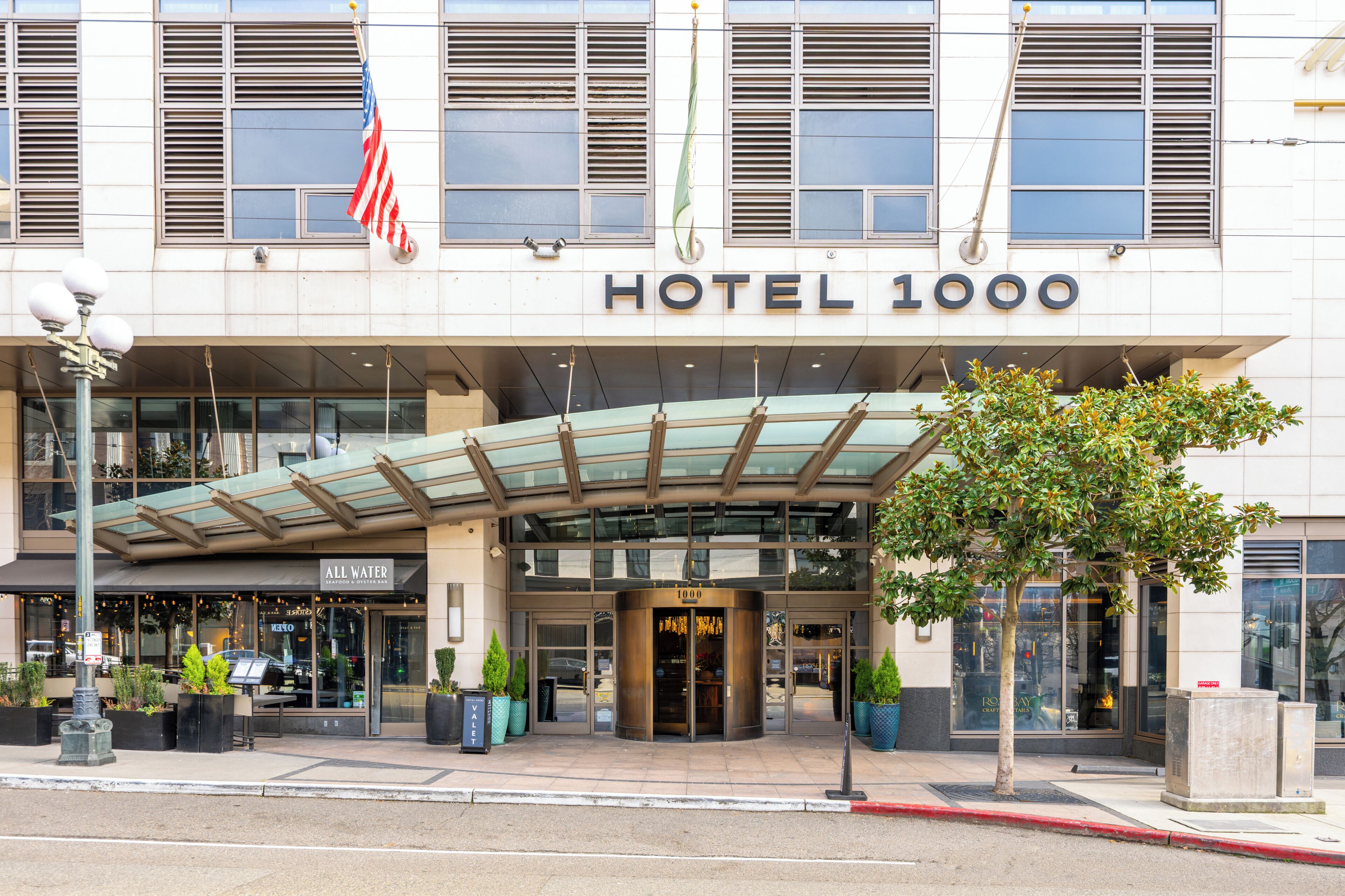 A hotel entrance with a glass canopy, an American flag, and a sign reading 'HOTEL 1000'.