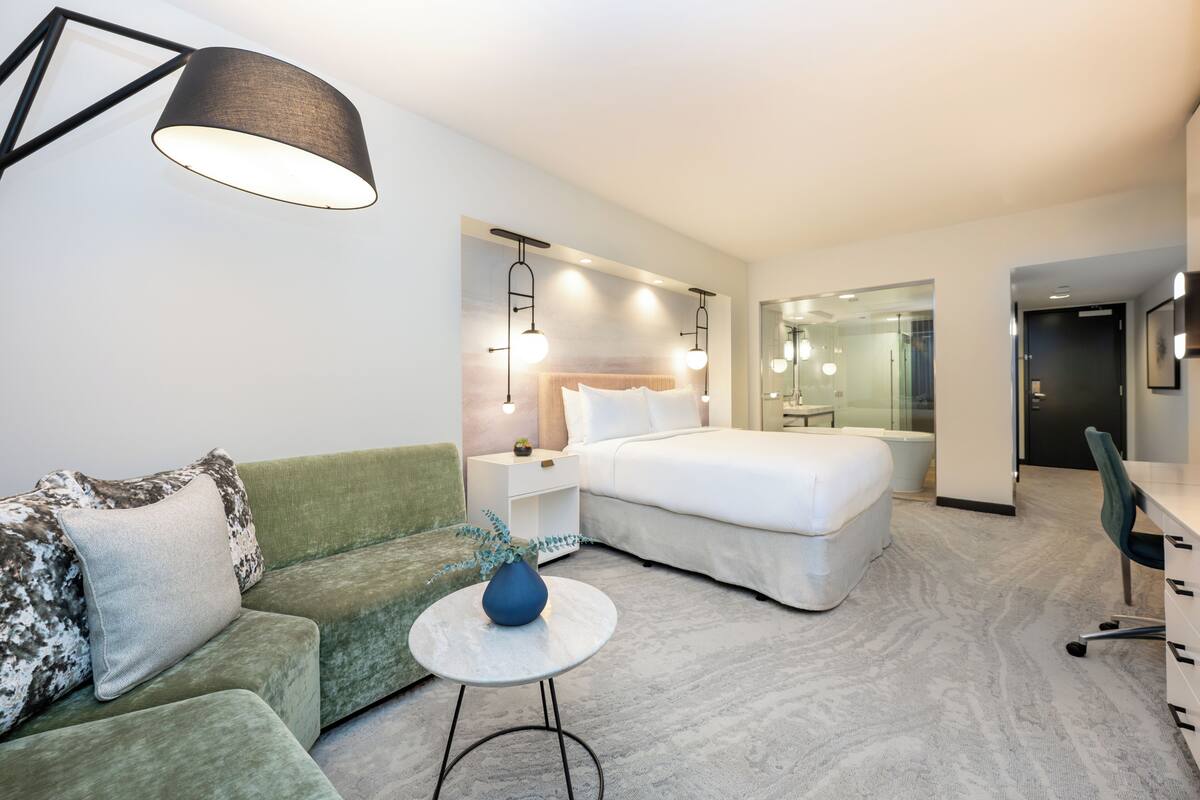 A modern hotel room with a green sofa, a white bed, a small round table with a vase, and a desk with a chair.