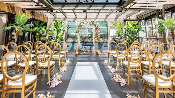 A glass-enclosed venue with rows of wooden chairs and a central aisle, surrounded by potted plants and a floral arrangement.