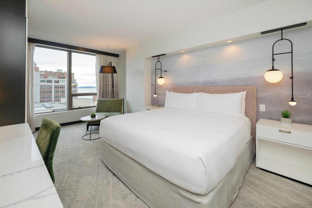A hotel room with a large bed, a desk, a chair, a small table, and a view of buildings through a large window.