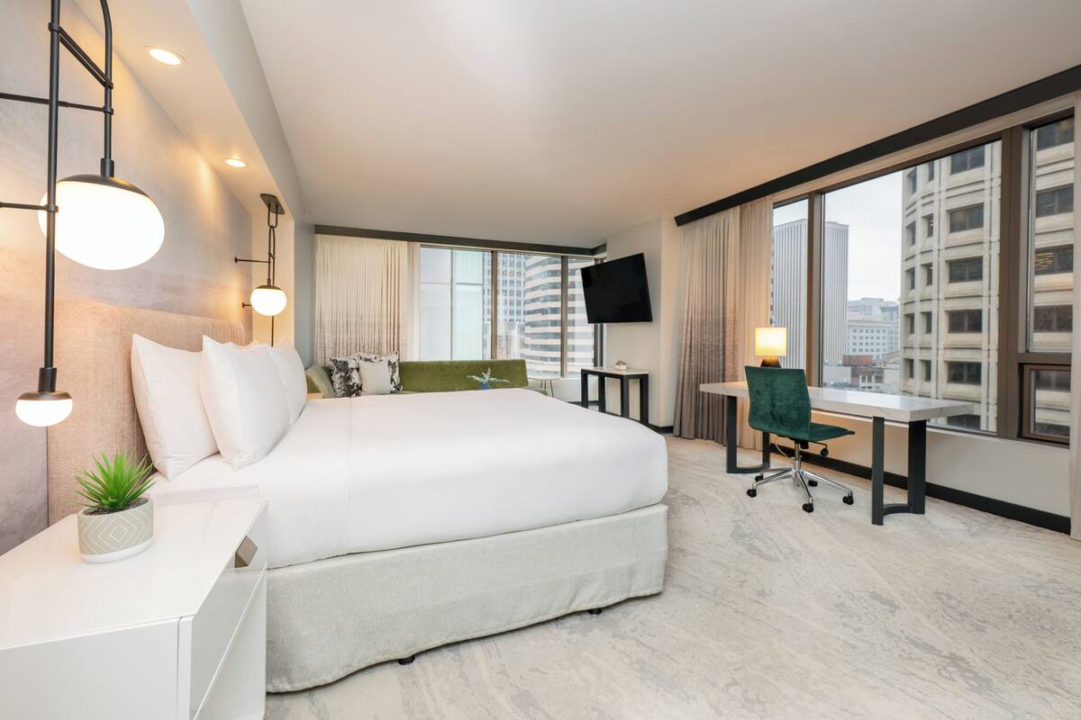 A modern hotel room with a large bed, a desk, a chair, a TV, and a city view.