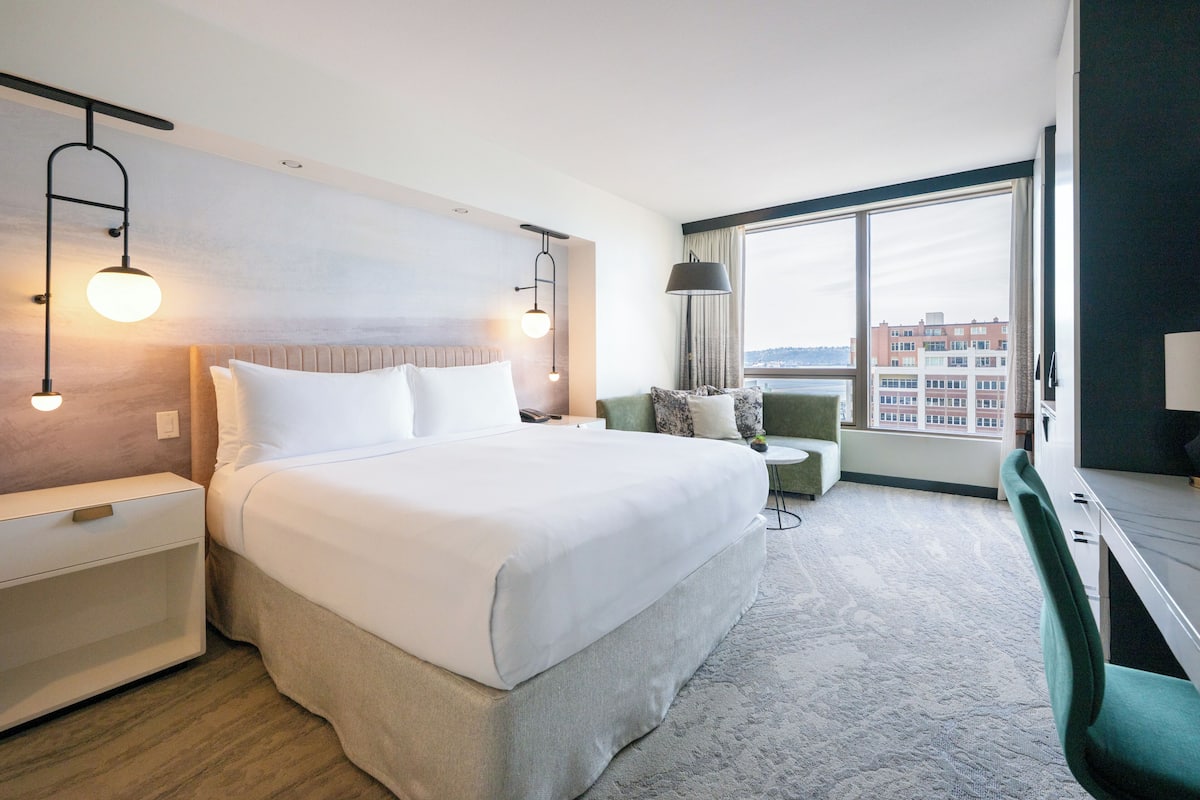 A hotel room with a large bed, bedside tables, a desk, a chair, and a view of buildings through large windows.
