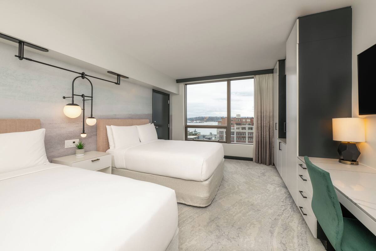 A hotel room with two beds, a desk, a TV, and a large window with a city view.