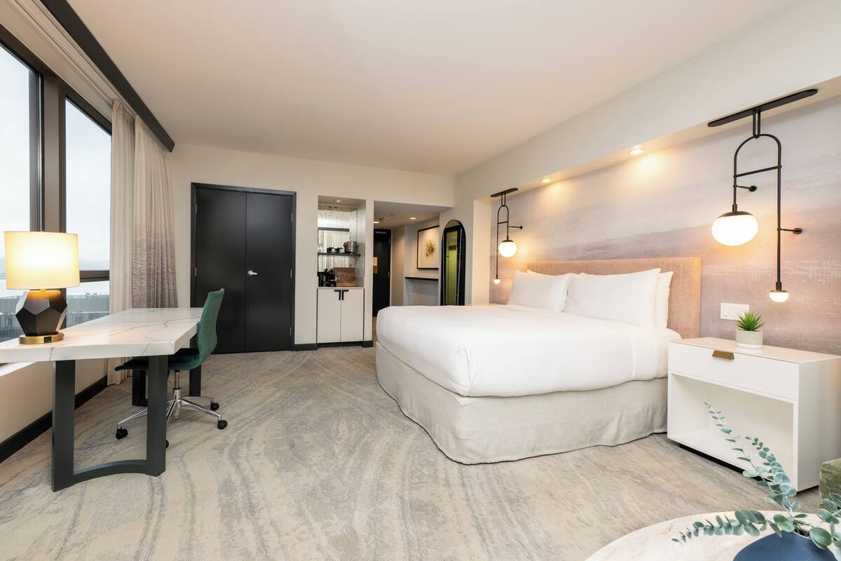 A hotel room with a bed, desk, chair, bedside table, lamp, and a view through the window.