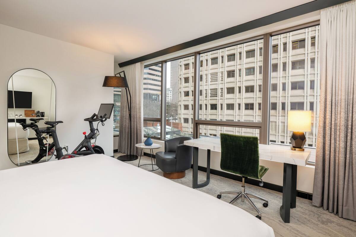 A hotel room with a bed, a desk, a chair, an exercise bike, a mirror, and large windows with a city view.