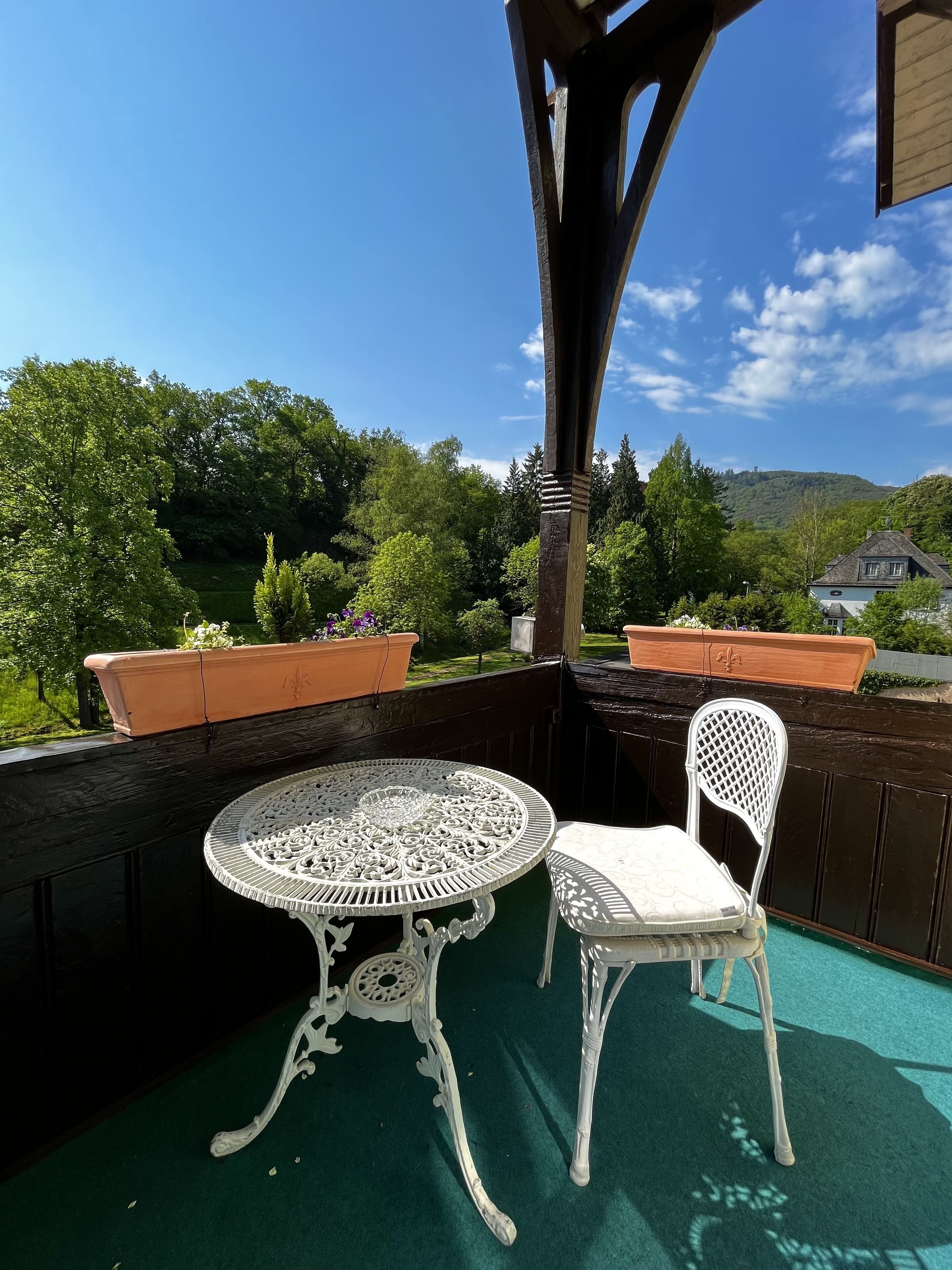 A covered outdoor area with a round table and a chair, surrounded by potted plants and a view of greenery and hills.
