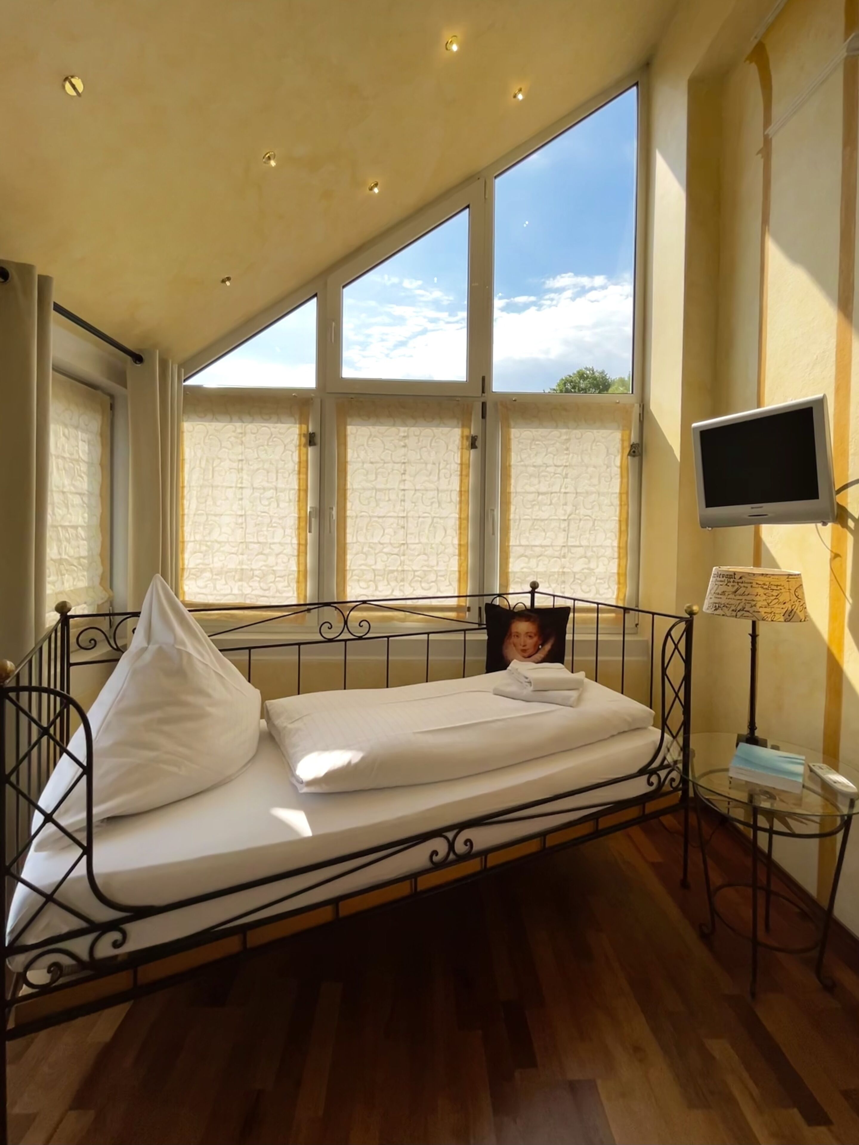Una cama individual con cabecera, una mesita de noche con lámpara, un televisor y una ventana con vistas al cielo.