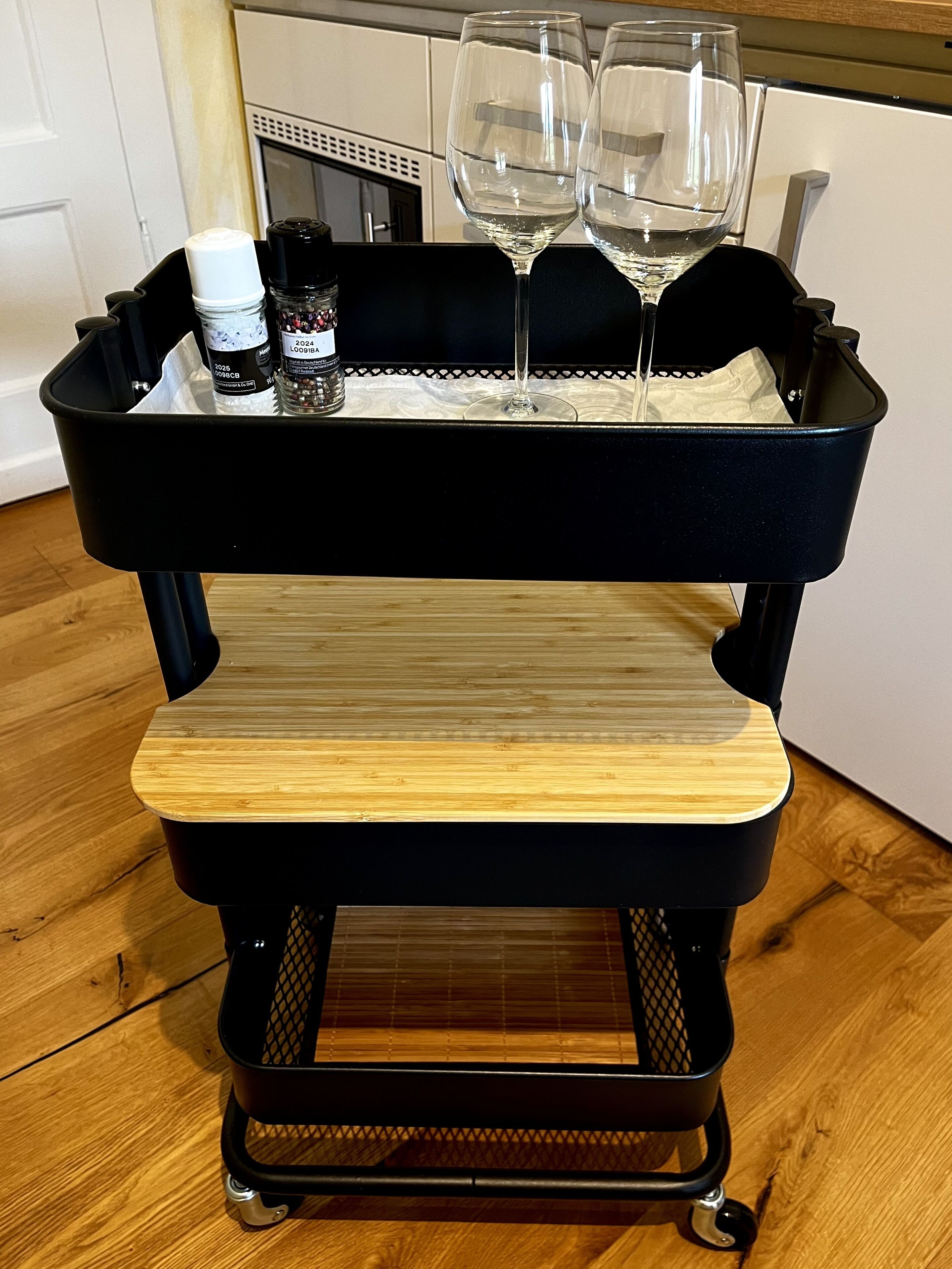 A black cart with a wooden top and wheels, containing two wine glasses and a bottle.
