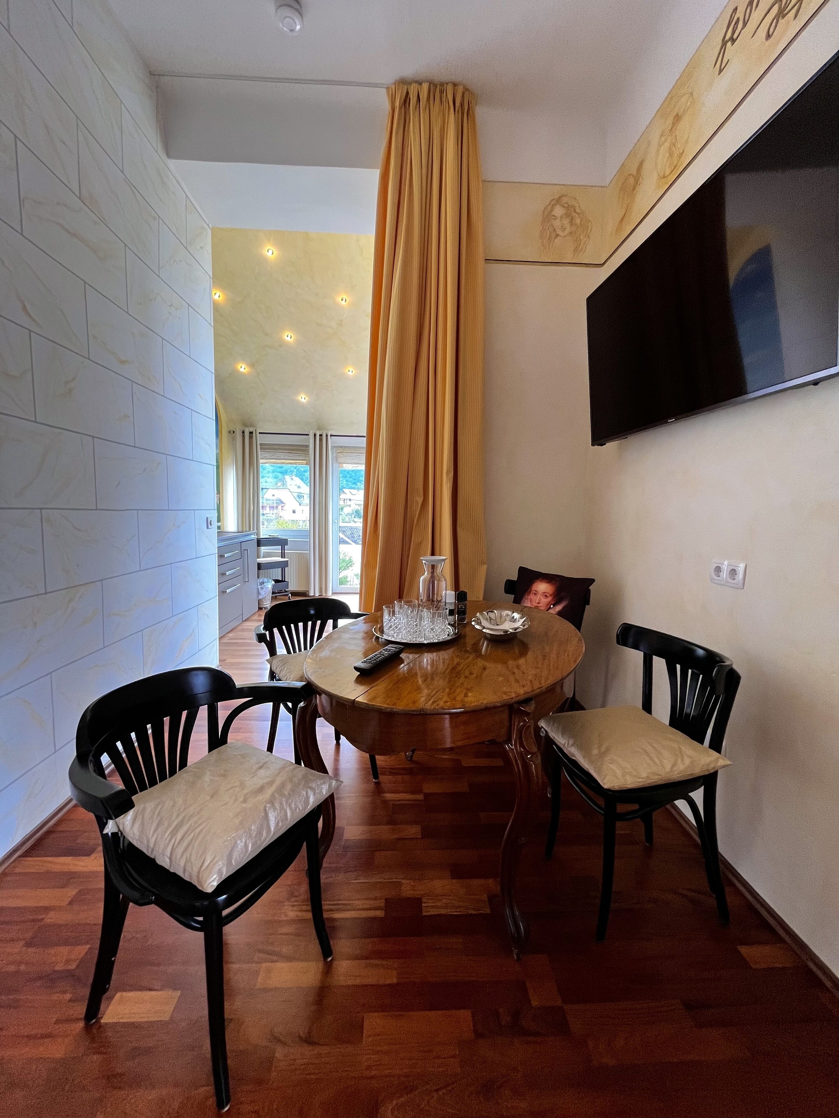 Un coin repas avec une table ronde en bois, des chaises noires dotées de coussins beige, un téléviseur fixé au mur et une grande fenêtre avec des rideaux.
