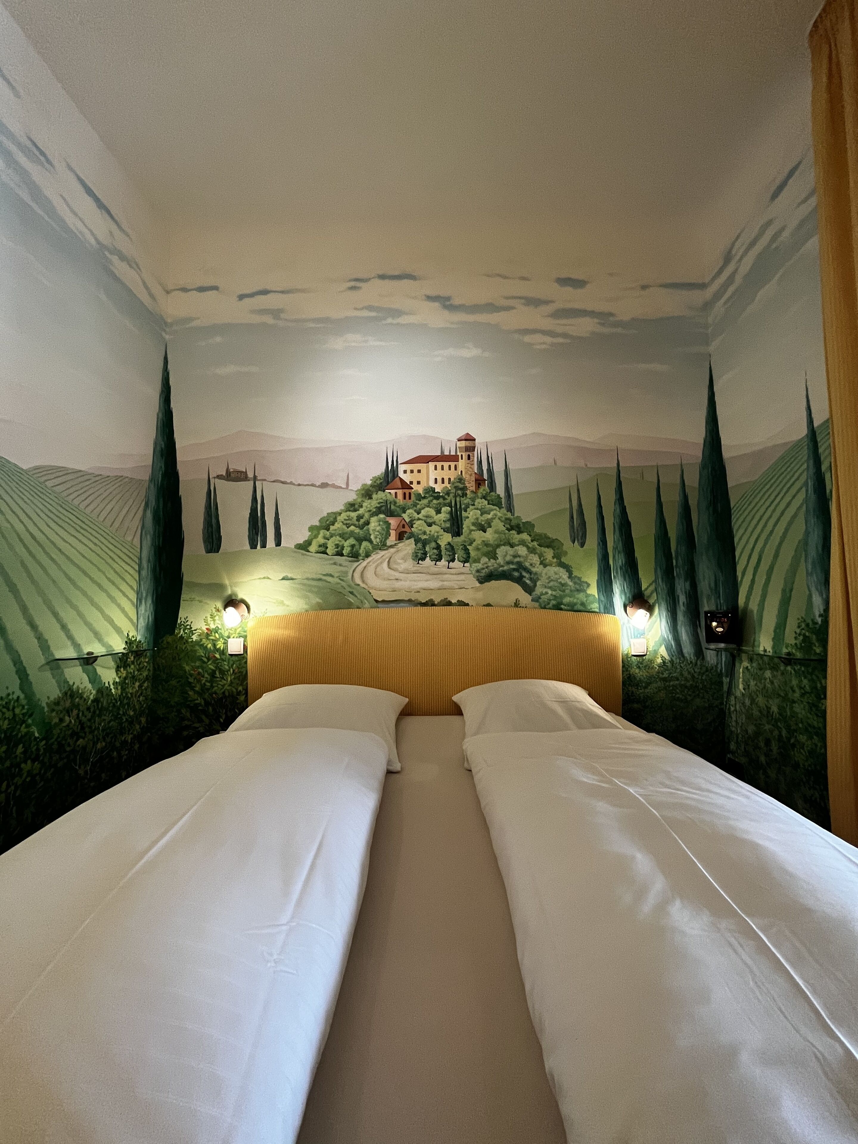 A room with a mural of a castle on the wall, a bed with white linens, and two bedside lamps.