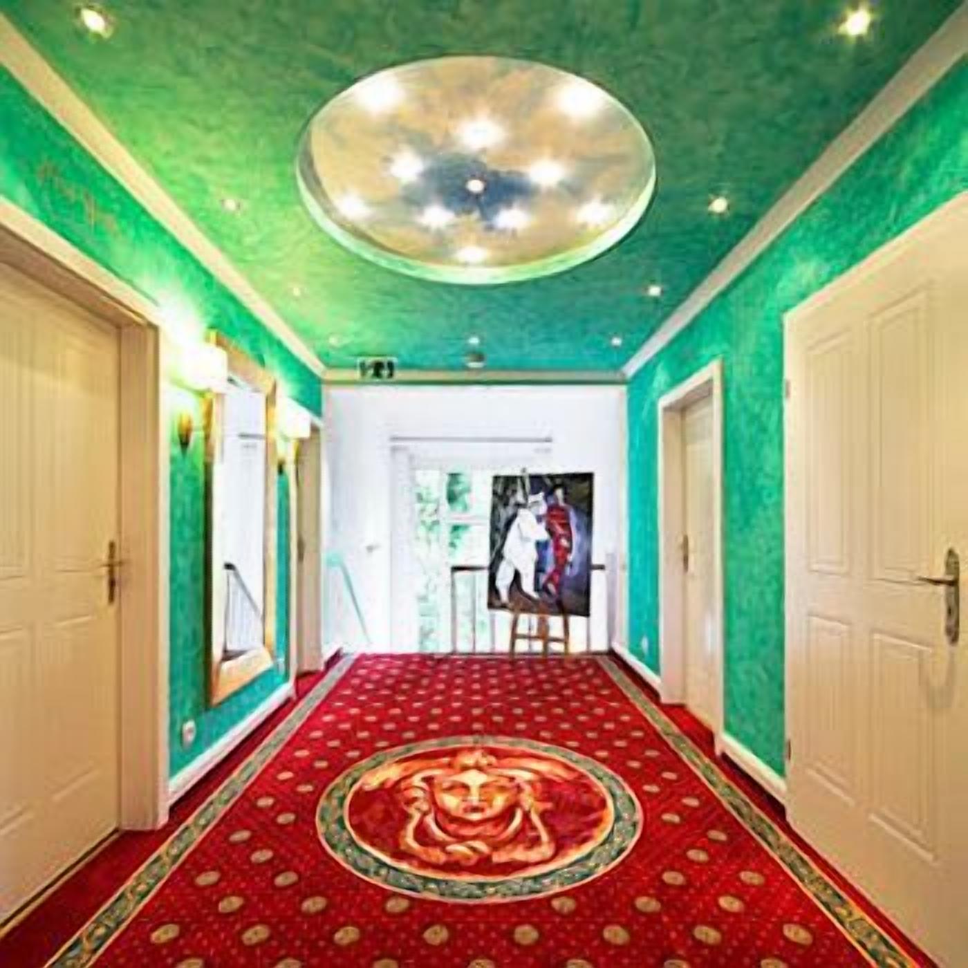 A hallway with a red carpet featuring a central emblem, white doors, and a green ceiling with a circular light fixture.