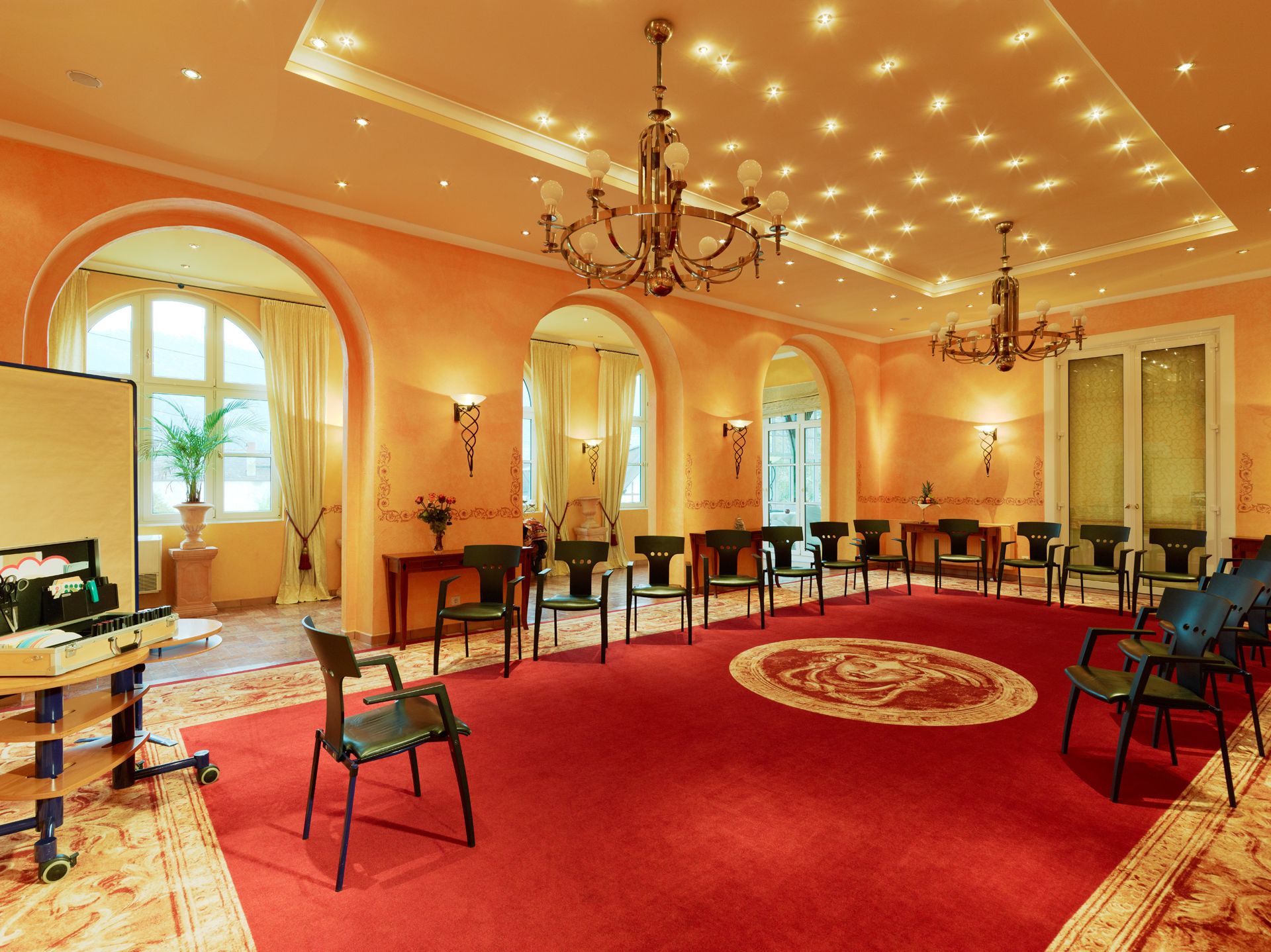 Une pièce spacieuse avec une moquette rouge, de nombreux fauteuils, un lustre et une grande fenêtre avec des rideaux.