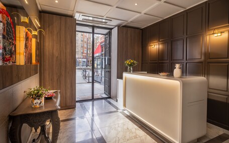 A modern hotel lobby with a reception desk, a wooden panel wall, and a view of the street outside.