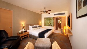 Quarto de hotel com cama, escrivaninha, cadeira, ventilador de teto e televisão.