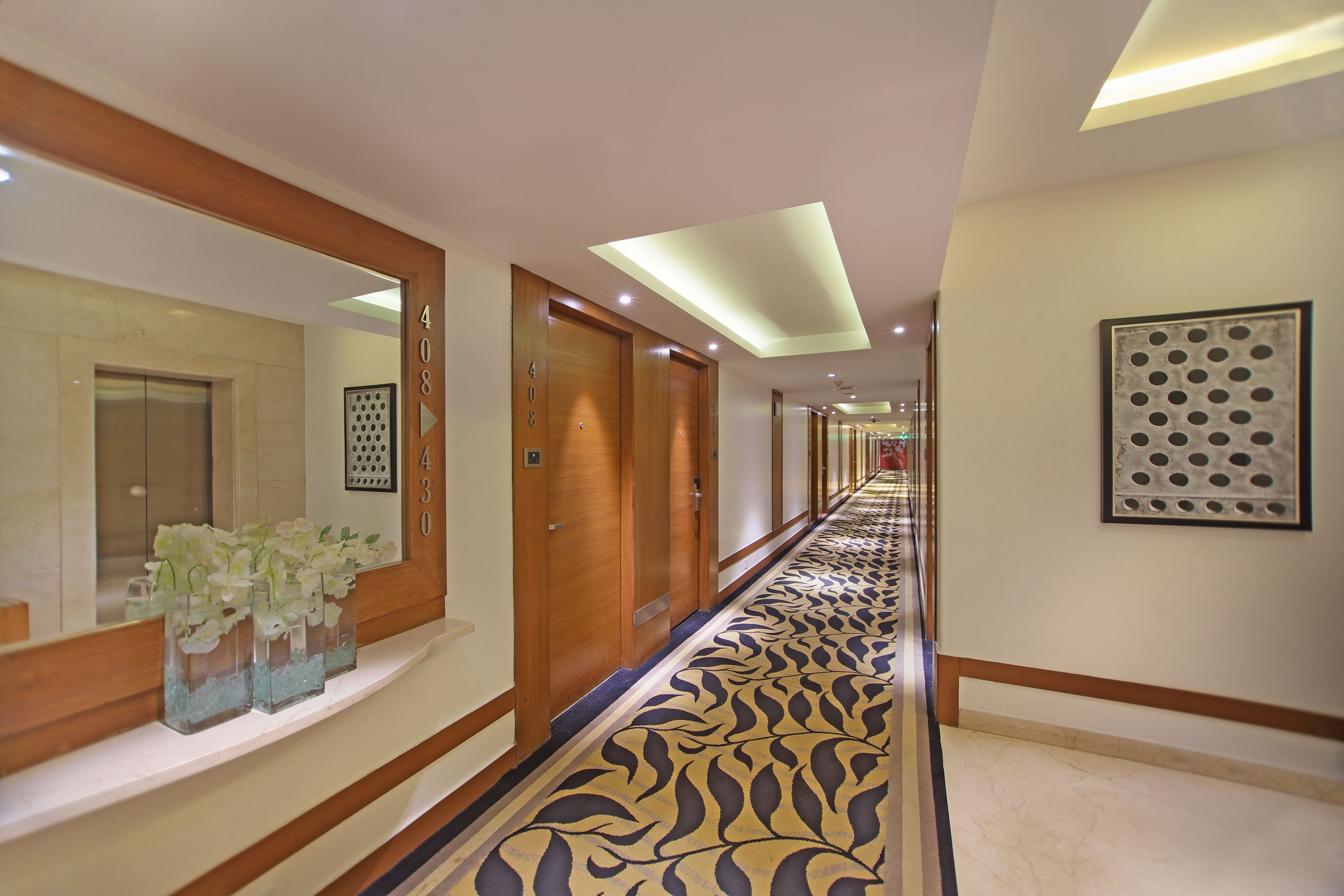 A hotel hallway with wooden doors, numbered rooms, a patterned carpet, and a mirror with a floral arrangement.