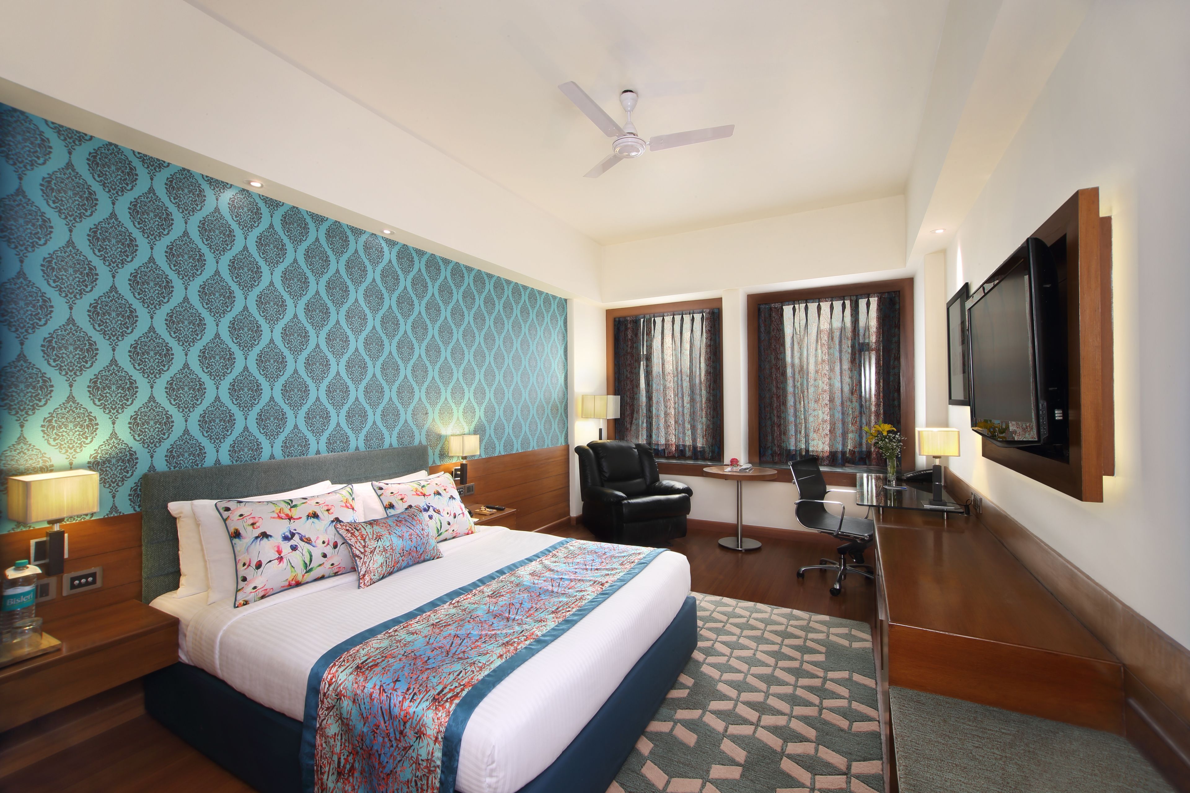 Une chambre d’hôtel avec un grand lit, deux tables de chevet, un bureau, une chaise, une télévision et un ventilateur de plafond.