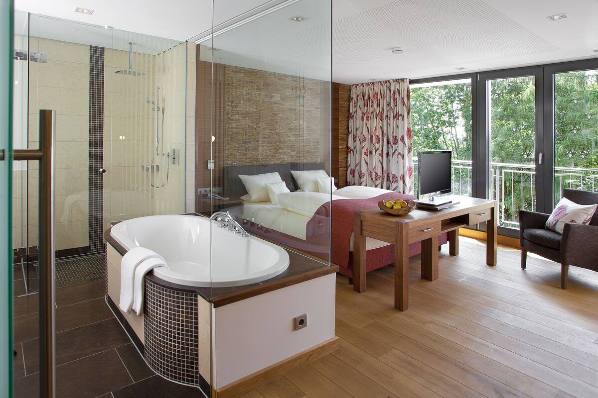 Ein modernes Hotelzimmer mit einer gläsernen Duschkabine, einer großzügigen Badewanne, einem Bett mit Holz-Kopfteil, einem Schreibtisch mit Computer und einem Balkon mit Blick auf Bäume.