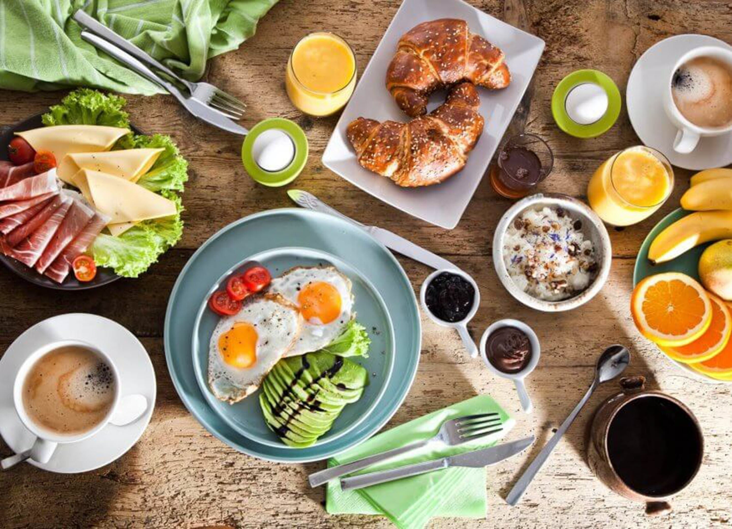 Une table en bois dressée pour le petit-déjeuner, avec des œufs, des croissants, du fromage et des fruits.