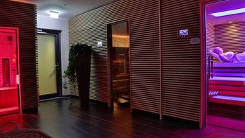 A modern hotel lobby with a wooden slatted wall, a reception desk, a plant, and a massage room with a purple-lit bed.