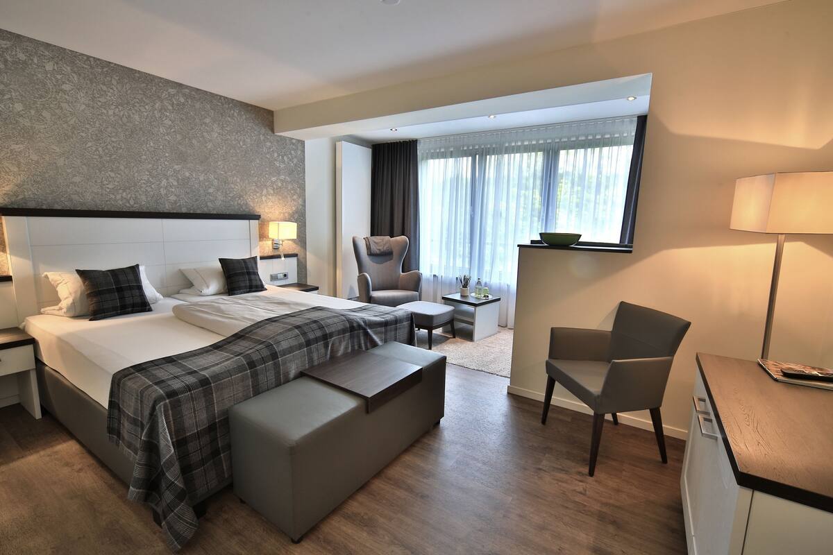 Ein modernes Hotelzimmer mit einem großen Bett, einem Schreibtisch, einem Sessel und Blick ins Freie.