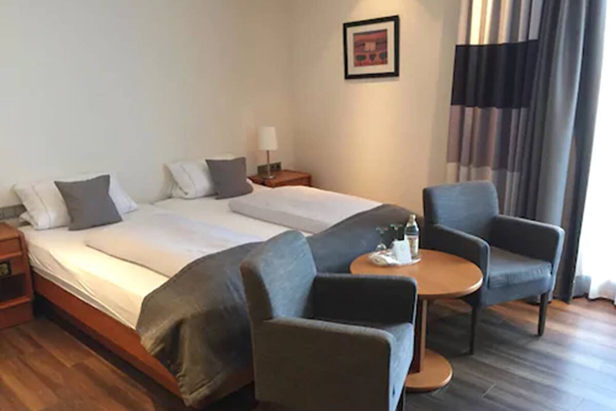 Ein Hotelzimmer mit zwei Betten, einem kleinen Tisch mit einer Flasche, zwei Sesseln, einem Nachttisch und einem gerahmten Bild an der Wand.