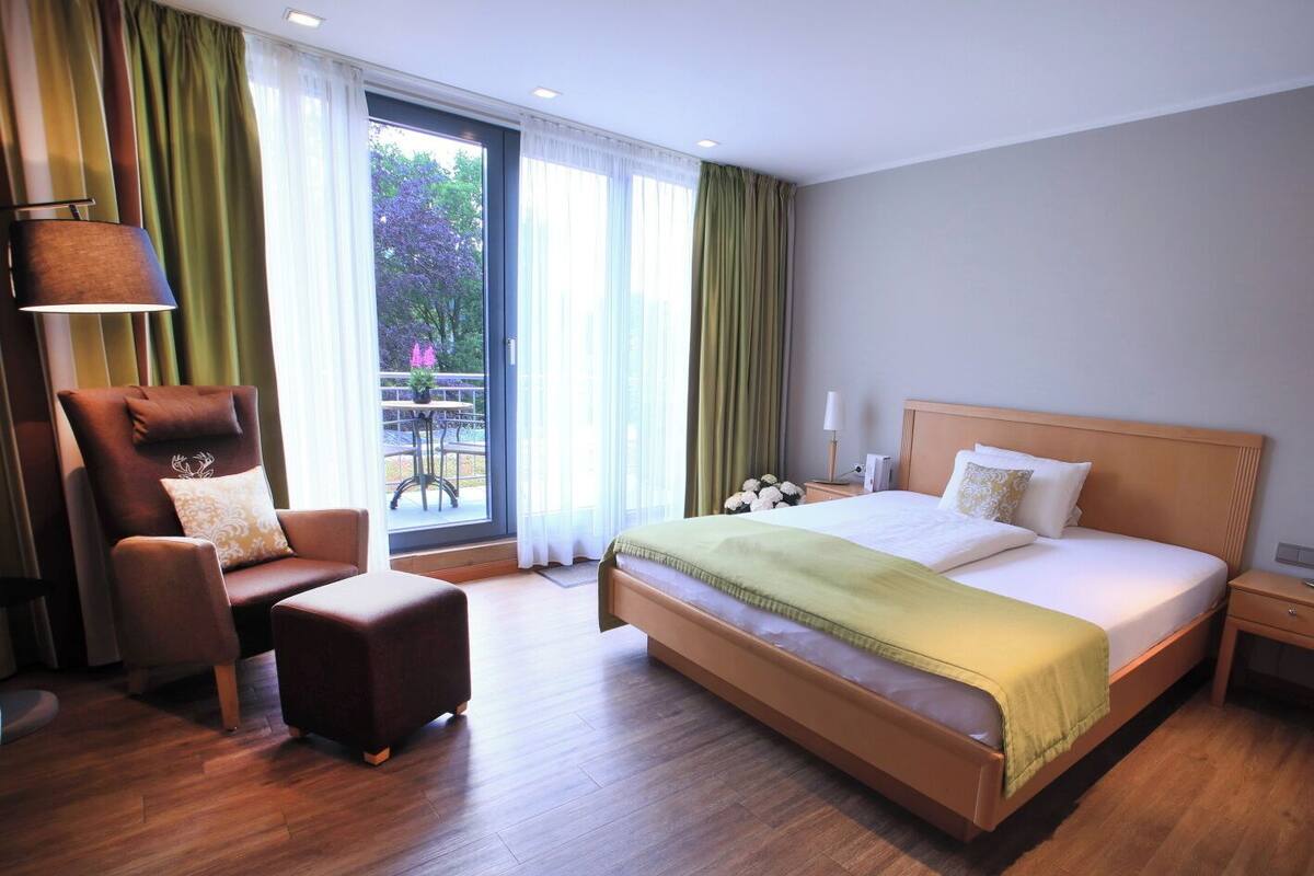 Ein Hotelzimmer mit einem großen Bett, einem Sessel, einem kleinen Tisch und Blick auf Grünfläche.