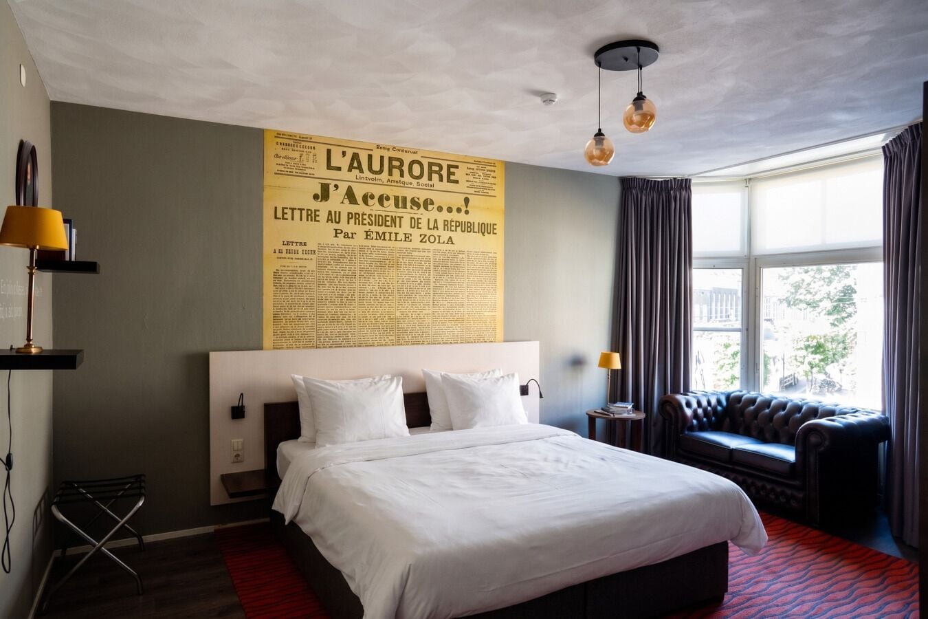 Hotelkamer met een groot bed, een bank, een raam met gordijnen en een muur met een krantenkoof.