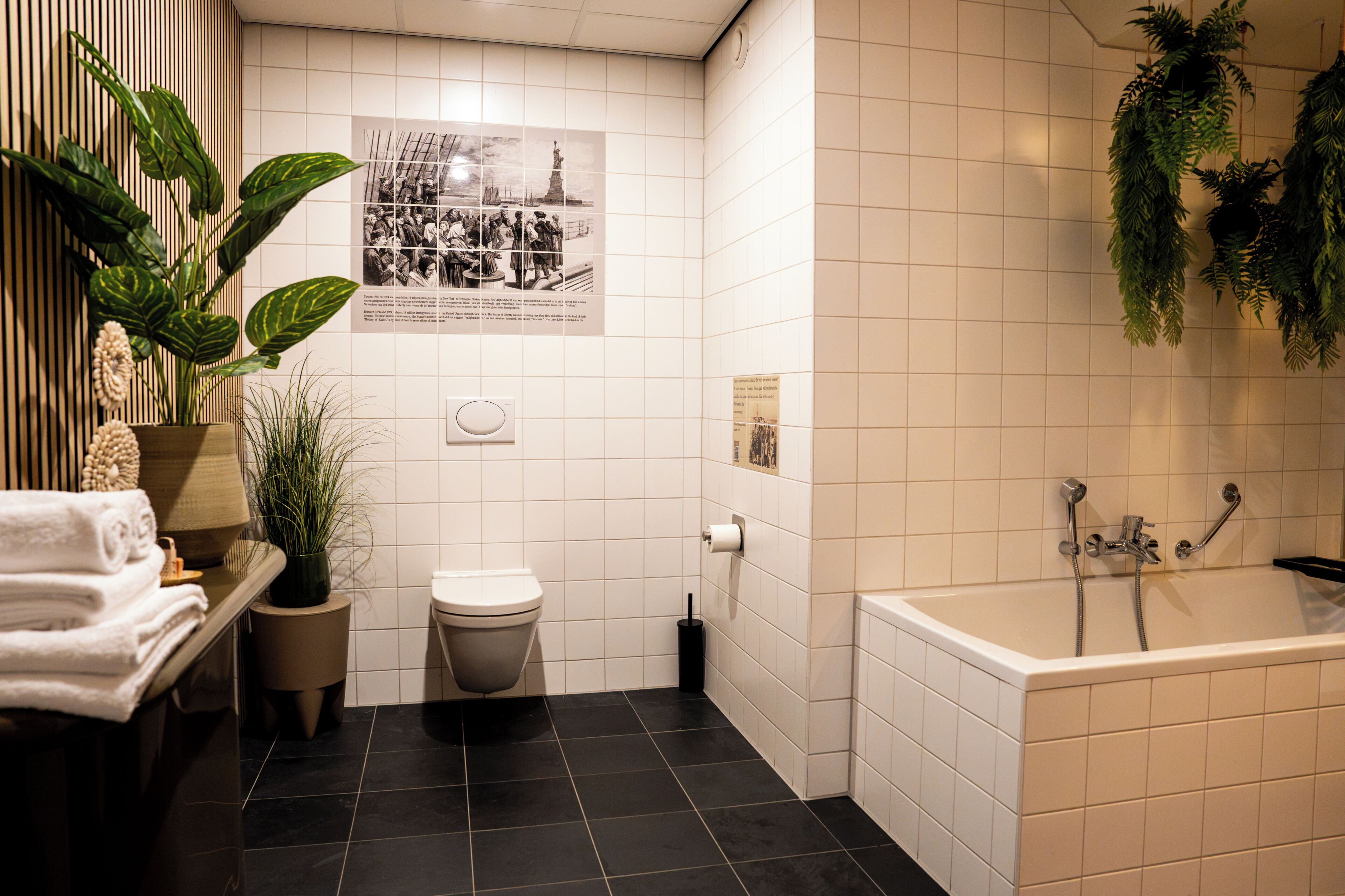 Une salle de bain avec une baignoire blanche, des toilettes et un lavabo. On y trouve également des plantes et une affiche accrochée au mur.