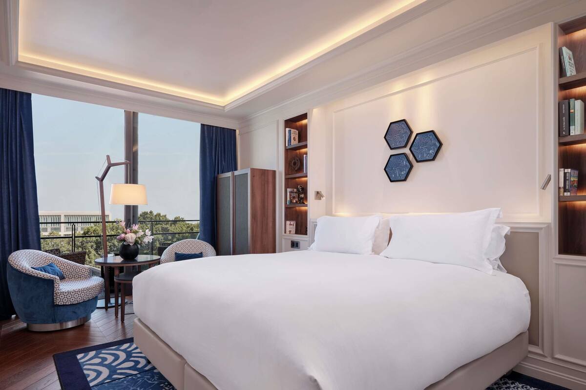 Ein Hotelzimmer mit einem großen Bett, einer Sitzecke mit Stuhl und Tisch, einem Bücherregal und Blick durch ein Fenster.