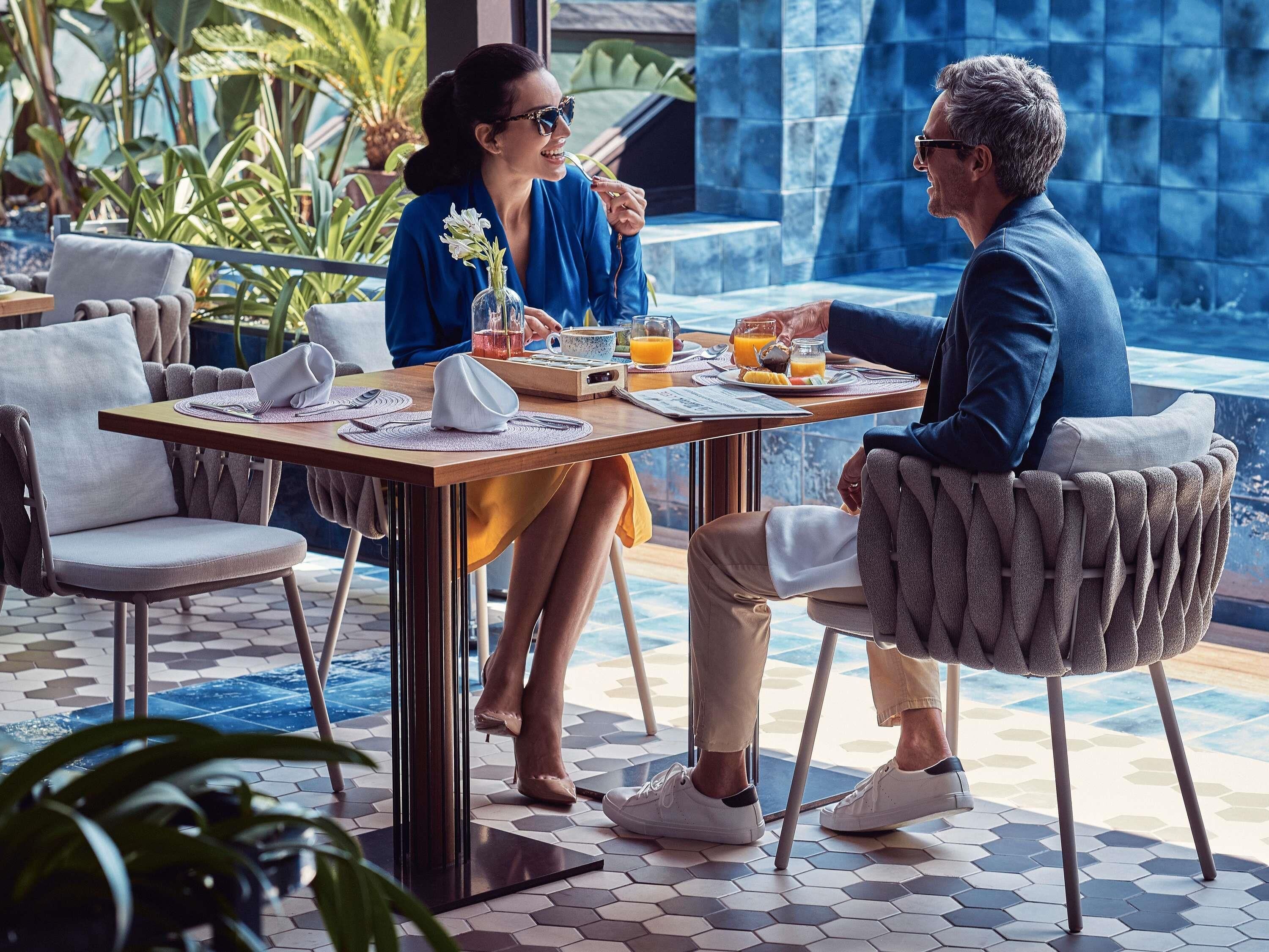 Un comedor con una mesa preparada para dos personas, rodeada de sillas con respaldo acolchado, suelo de baldosas y vistas a una fuente.