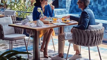 Un comedor con una mesa preparada para dos personas, rodeada de sillas con respaldo acolchado, suelo de baldosas y vistas a una fuente.