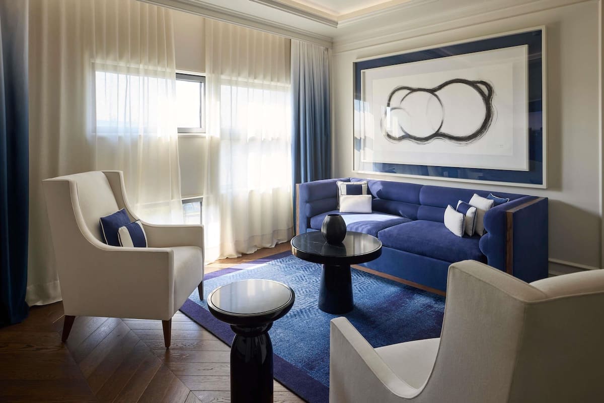 Ein modernes Wohnzimmer mit einem blauen Sofa, weißen Sesseln, einem runden Couchtisch und einem großen abstrakten Gemälde an der Wand.
