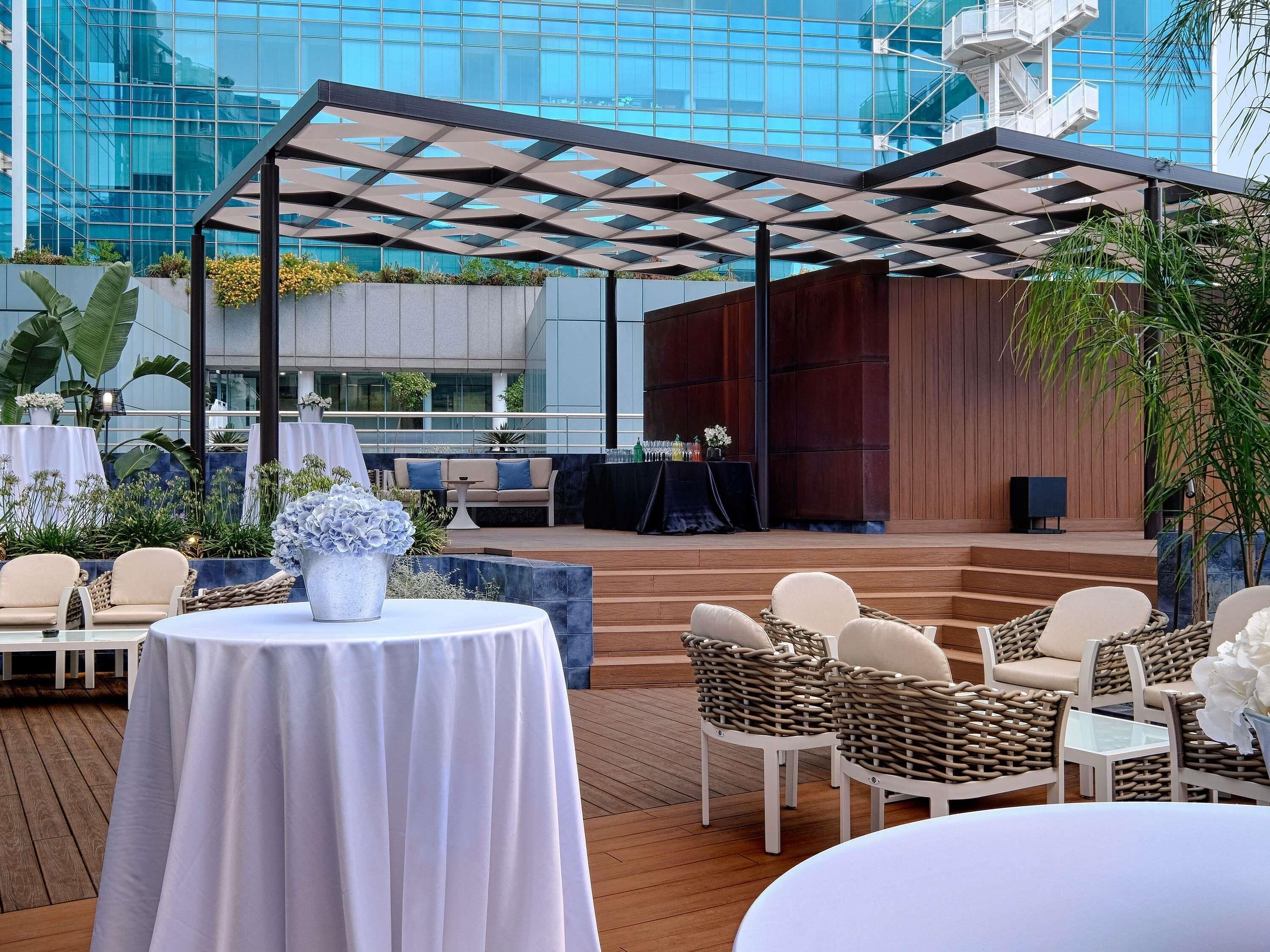An outdoor patio with tables and chairs, a stage, and a modern building in the background.