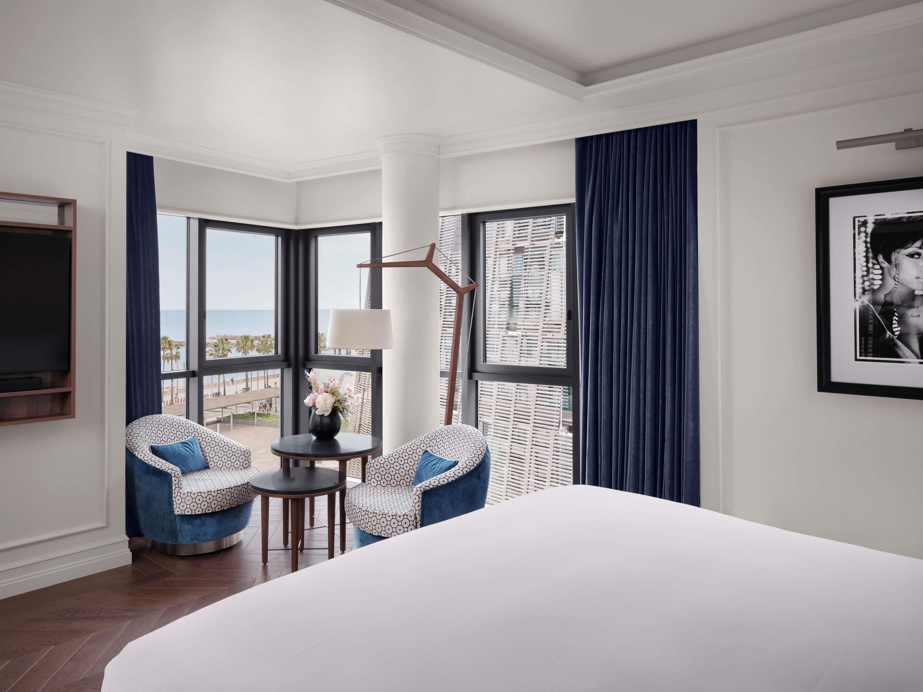 Quarto de hotel com uma cama grande, duas poltronas estampadas, uma mesinha com abajur, um quadro emoldurado e vista para a praia.