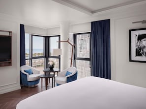 A hotel room with a large bed, two patterned armchairs, a small table with a lamp, a framed picture, and a view of the beach.