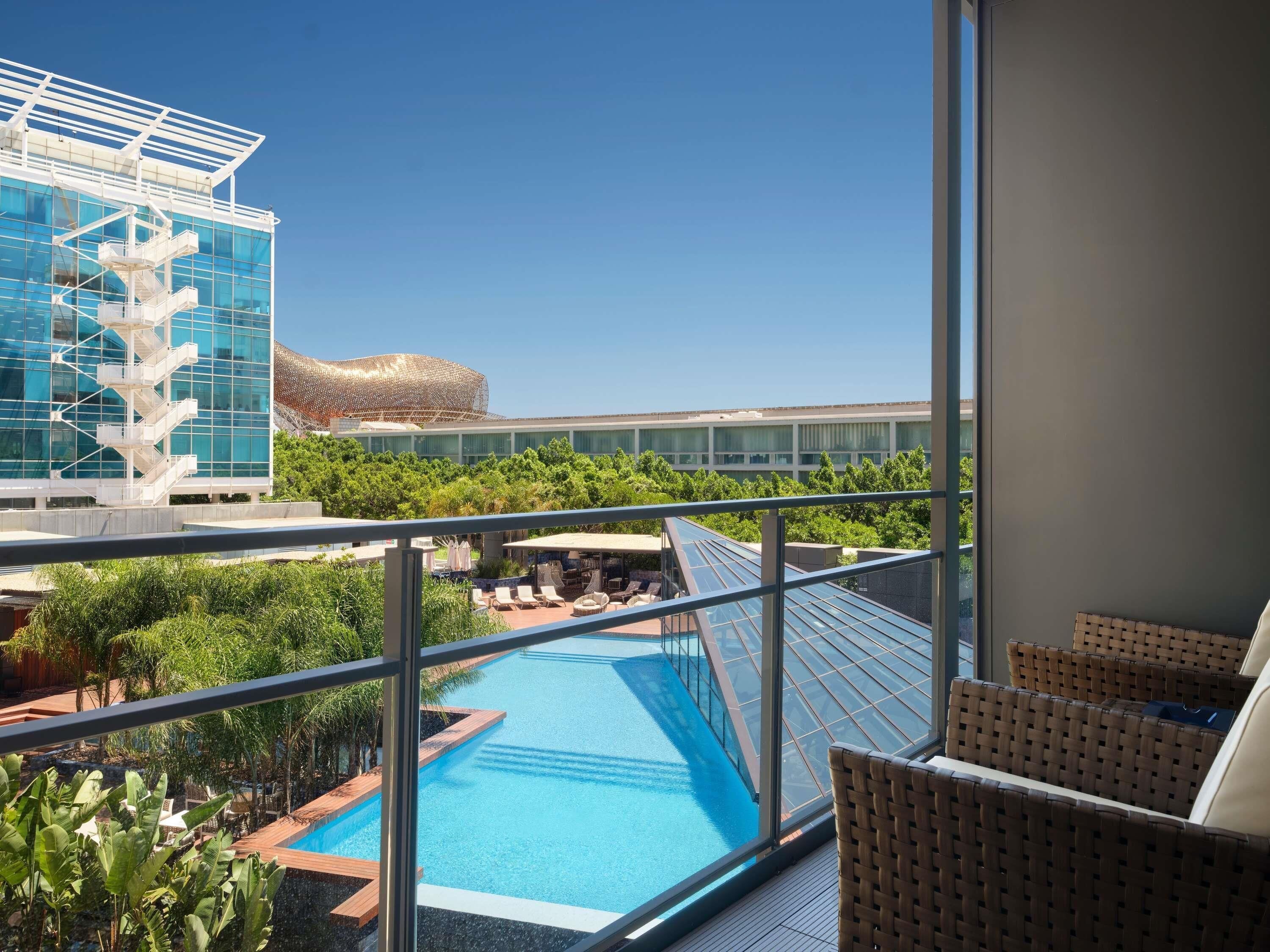 A poolside view with a glass balustrade, lounge chairs, and a modern building with a unique roof structure.