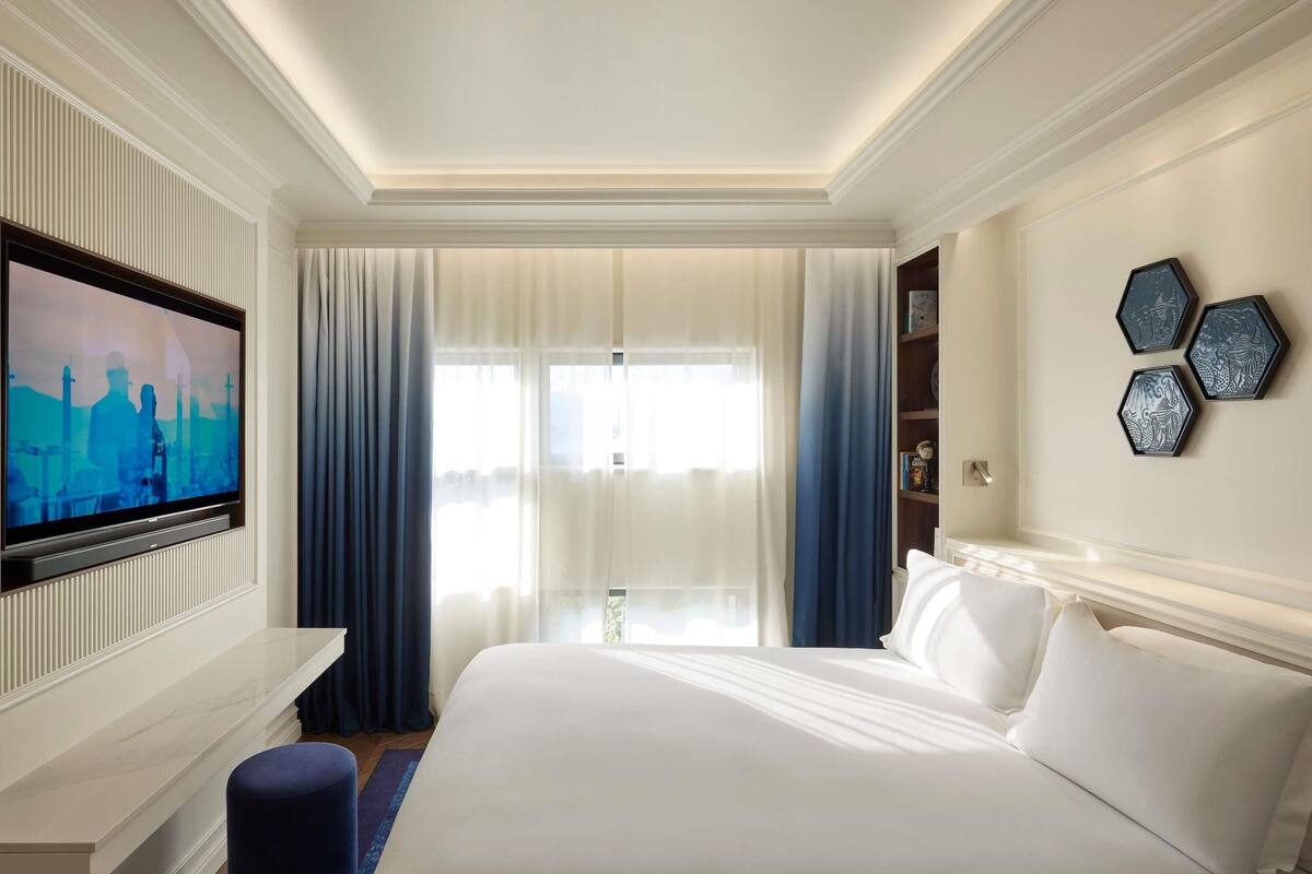 A hotel room with a large bed, a flat-screen TV mounted on the wall, a bookshelf, and a window with curtains.