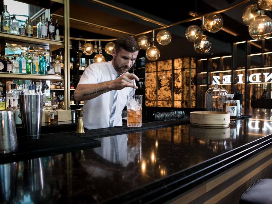 A bartender preparing a drink at a bar with shelves of liquor bottles and a polished counter.
