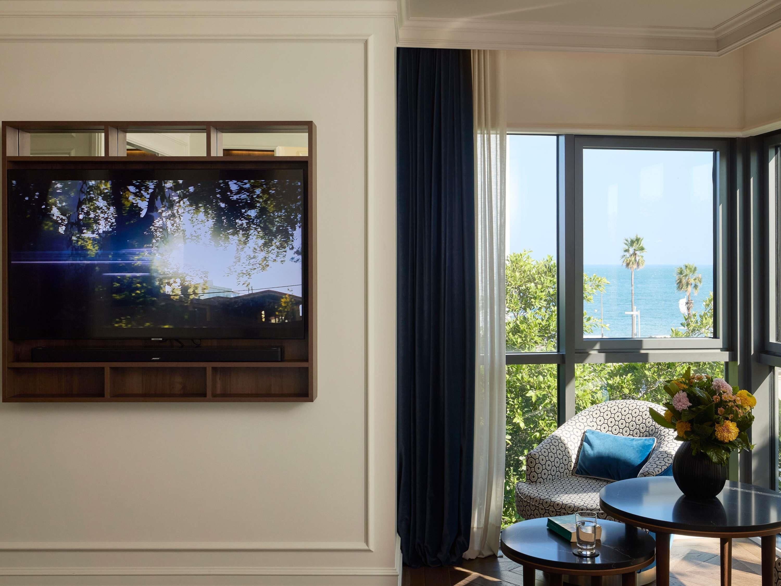 A room with a television mounted on the wall, a window with a view of the sea, a sofa with patterned cushions, and a small table with a vase of flowers.