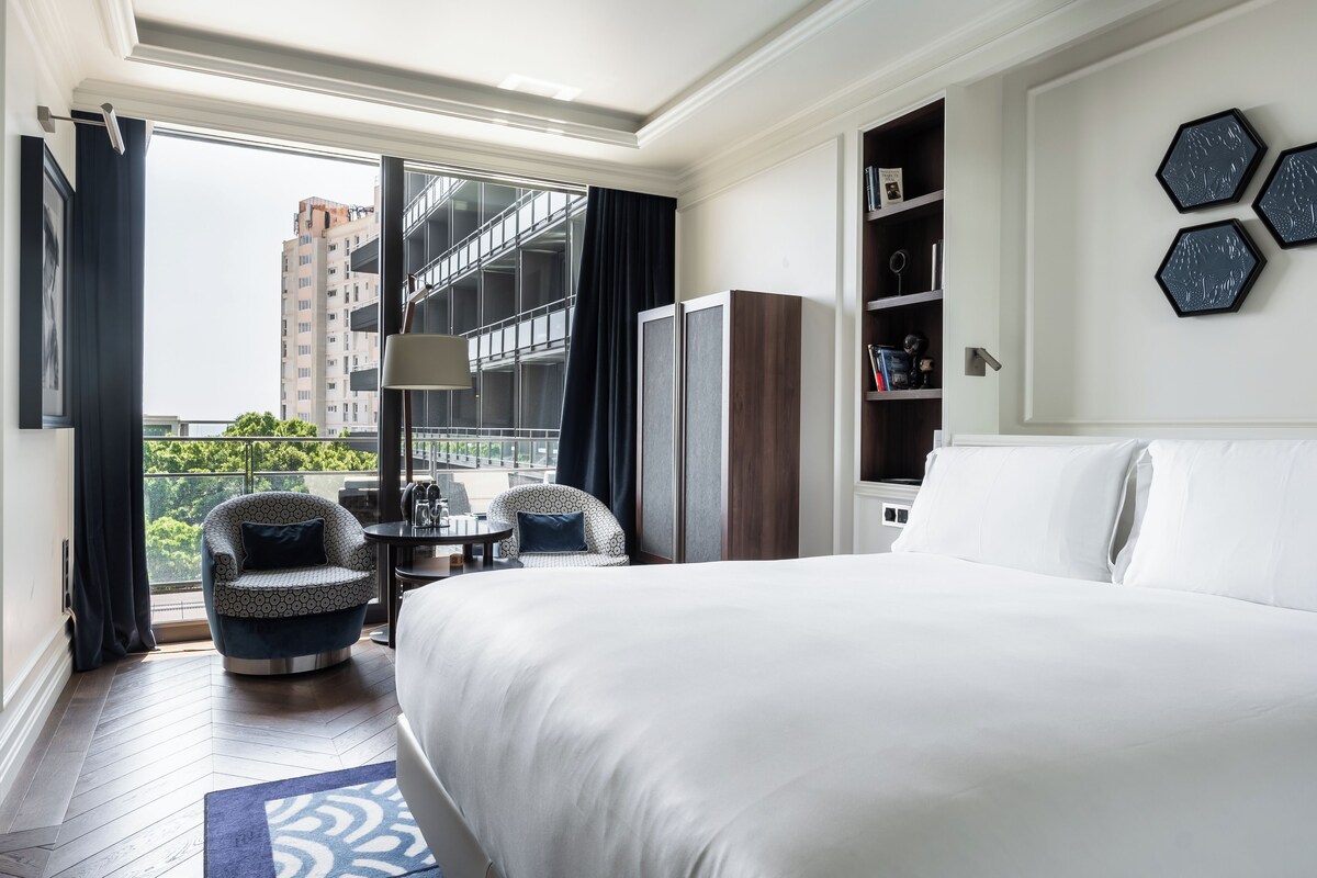 A hotel room with a large bed, two armchairs, a small table, and a bookshelf. There is a balcony with a view of a building and greenery.