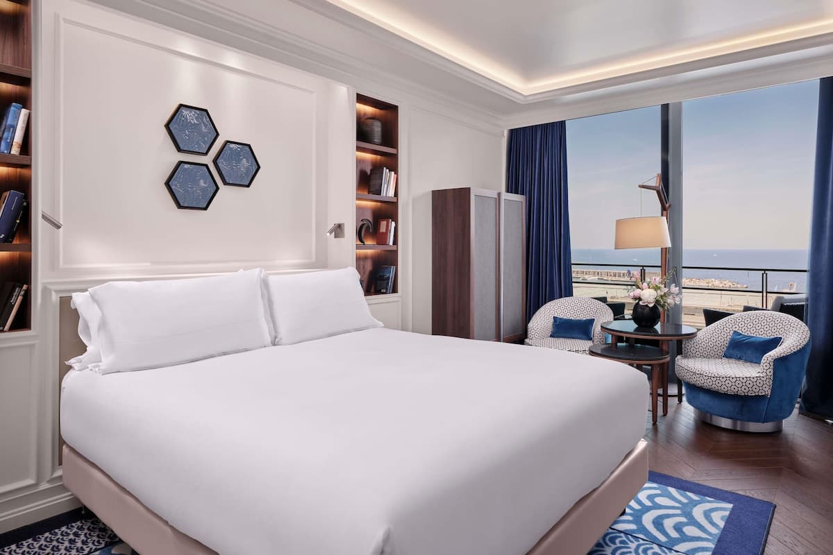 A hotel room with a large bed, a seating area with two chairs, a bookshelf, and a view of the sea.