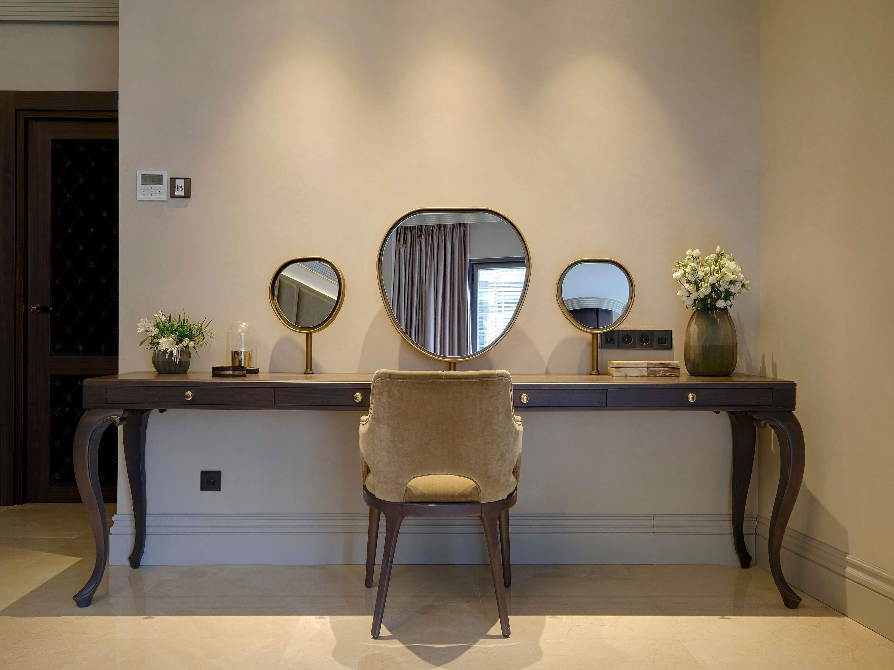 Un escritorio con tres espejos, un florero con flores y una silla en una habitación de hotel.